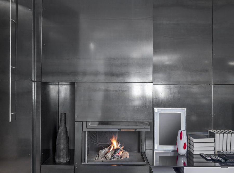 Interiors of  a modern living room in the foreground the fireplace integrated on the wall covered of a iron panels the floor is made of wood